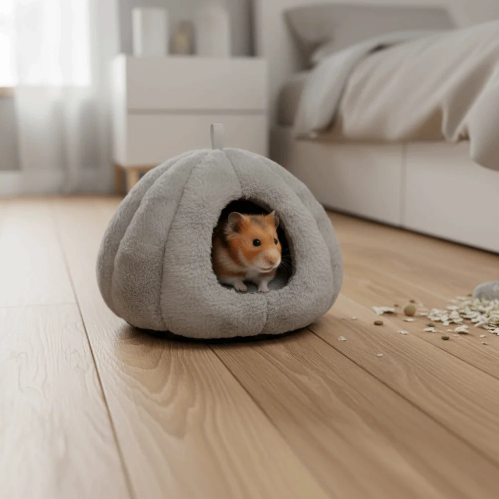 A small orange hamster peeks out of a gray plush hanging nest on a wooden floor.