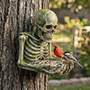 A sculpted skull and skeleton figure made of resin is attached to a tree trunk, serving as an outdoor garden decoration.