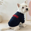 A white dog wearing a navy blue hoodie with red text on the back, sitting on a light surface.