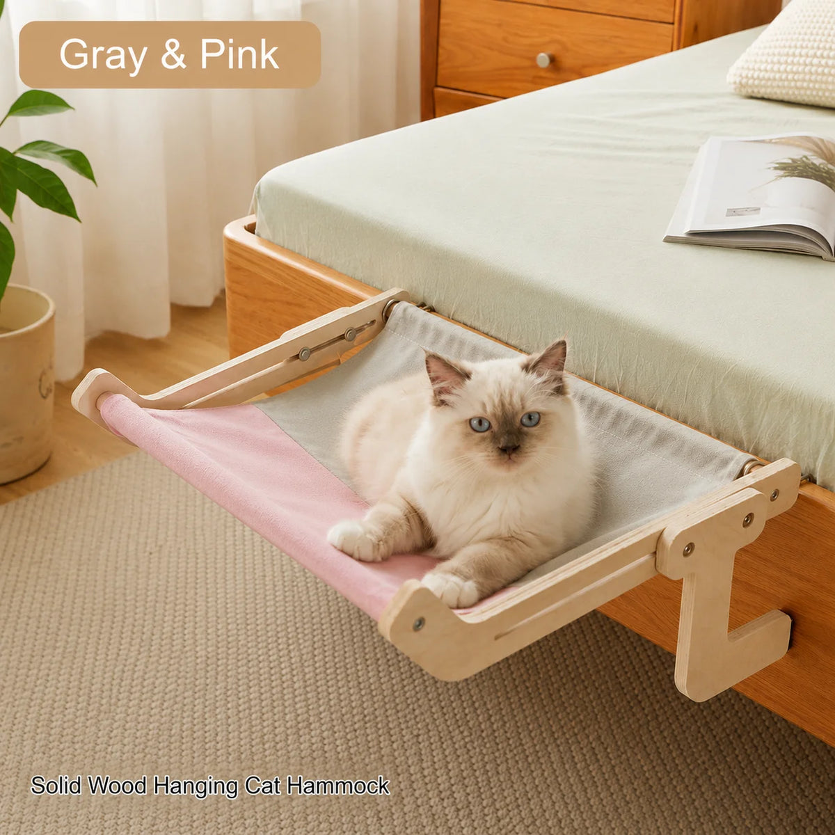 A solid wood hanging cat hammock in pink and gray fabric resting on a bed.
