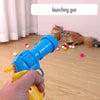 Eine Hand hält eine blaue Startpistolen-Spielzeugwaffe auf eine Katze und bunte Bälle auf einem Holzfußboden.