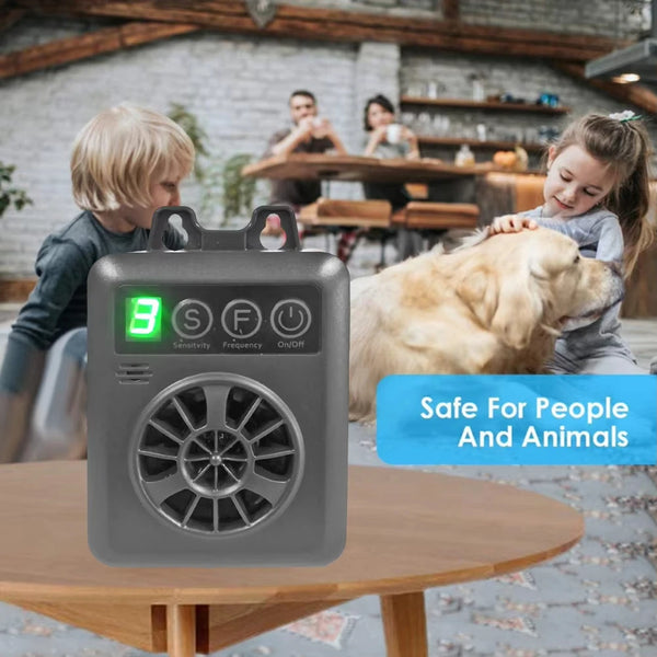 A gray ultrasonic anti-barking device is shown on a wooden table with a dog and people in the background.