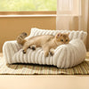 Un chaton roux et duvete relaxe dans un lit canapé beige texturé sur un sol clair.