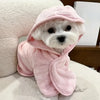 A white dog wearing a pink absorbent quick-dry bathrobe with a hood, resting on a white surface.