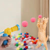 Eine Hand hält eine gelbe Spielzeugwaffe, die rosa und grüne Plüschbälle auf eine orange Katze feuert.
