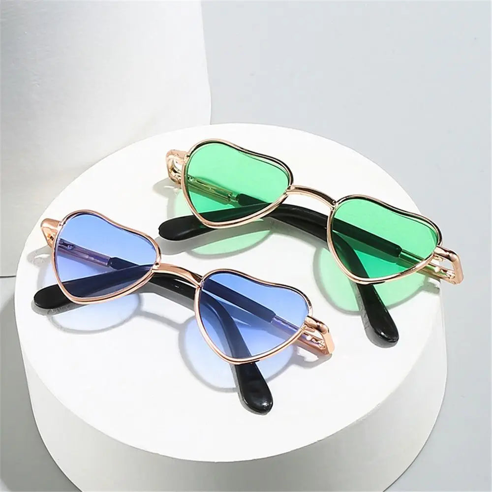 Pair of heart-shaped pet sunglasses with green and blue lenses displayed on a white stand.