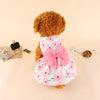 A fluffy brown dog wearing a pink floral bow dress with white trim, lying on a light background.