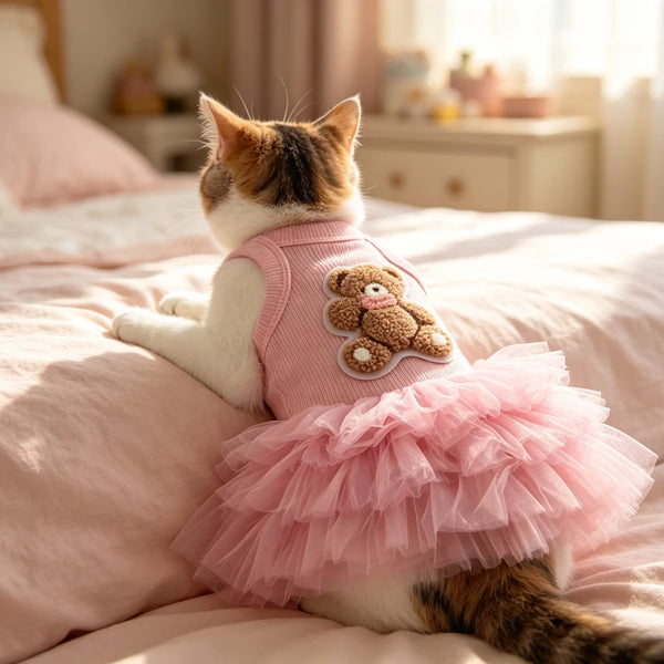 A fluffy tabby cat wearing a pink ruffled skirt with a teddy bear appliqué, lying on a pink blanket.