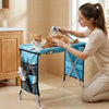 A woman is using a foldable pet grooming hammock to bathe and trim a cat in a kitchen setting.