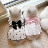 A fluffy white cat wearing a pink and black polka dot summer dress with a bow, lying on a light-colored surface.