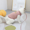 A white cat wearing a pink knitted t-shirt with a small tan patch, sitting on a light surface.
