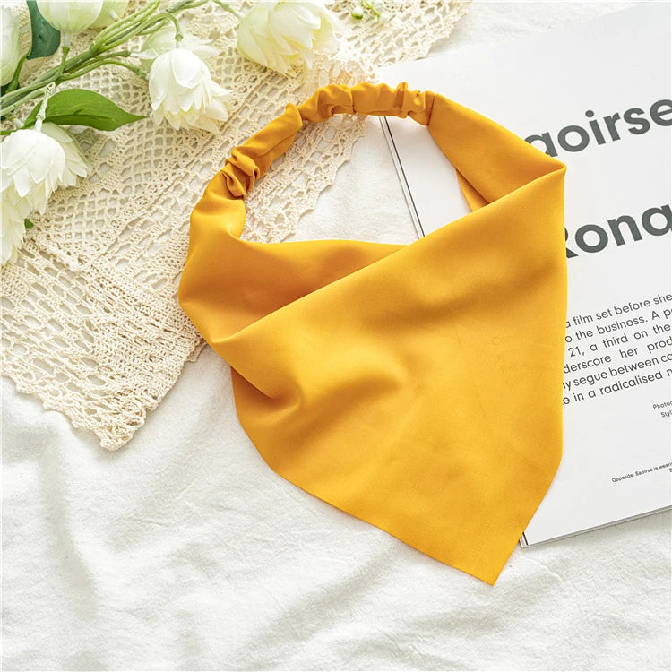 A bright yellow triangle scarf is draped on a white surface next to a white lace textile and some flowers.