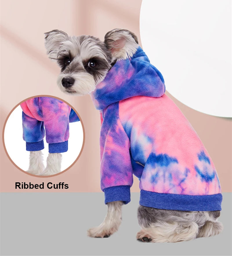 A small dog wearing a tie-dye fleece hoodie with ribbed cuffs and a purple and pink color pattern.