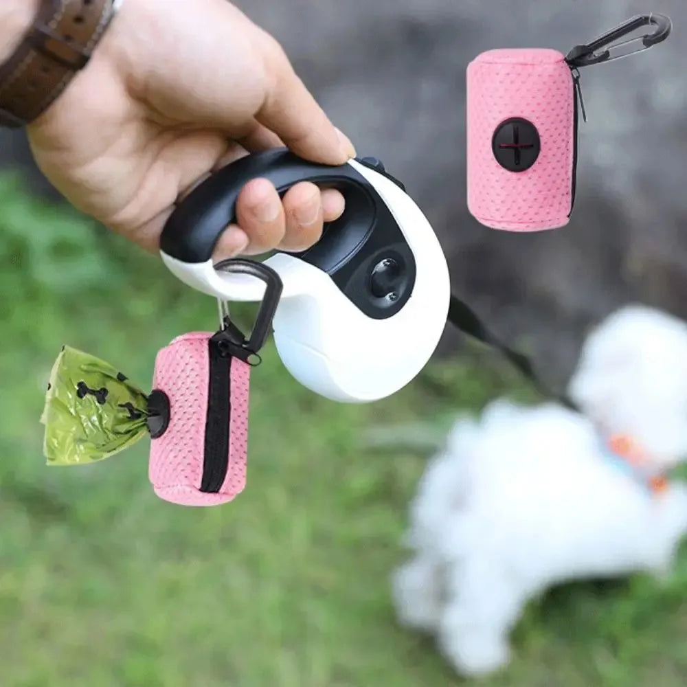 A hand holds a white and black portable mesh dog waste bag dispenser with a pink bag attached, against a green outdoor backgr