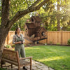 A woman is installing a rustic wooden bird house in a backyard setting under a tree.