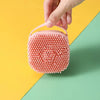 A hand is holding a pink silicone pet bathing massage glove with a textured, bubbly surface against a yellow and green backgr