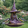 A dark purple resin witch hat sculpture with long hair sits on a shallow bowl, surrounded by small birds in an outdoor garden