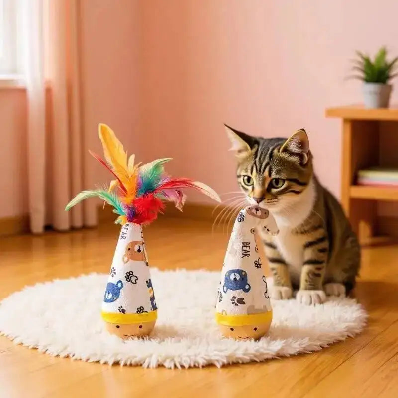 A tabby cat is interacting with a white and yellow feather wobble ball toy on a fluffy rug.