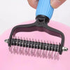 A hand holds a blue and black pet detangling comb with stainless steel teeth against a pink background.