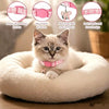 A fluffy gray kitten wearing a pink aesthetic cat collar with a bow pendant resting on a white pet bed.