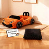 An orange supercar cat bed with black accents and a separate black plush cushion on a wooden floor.