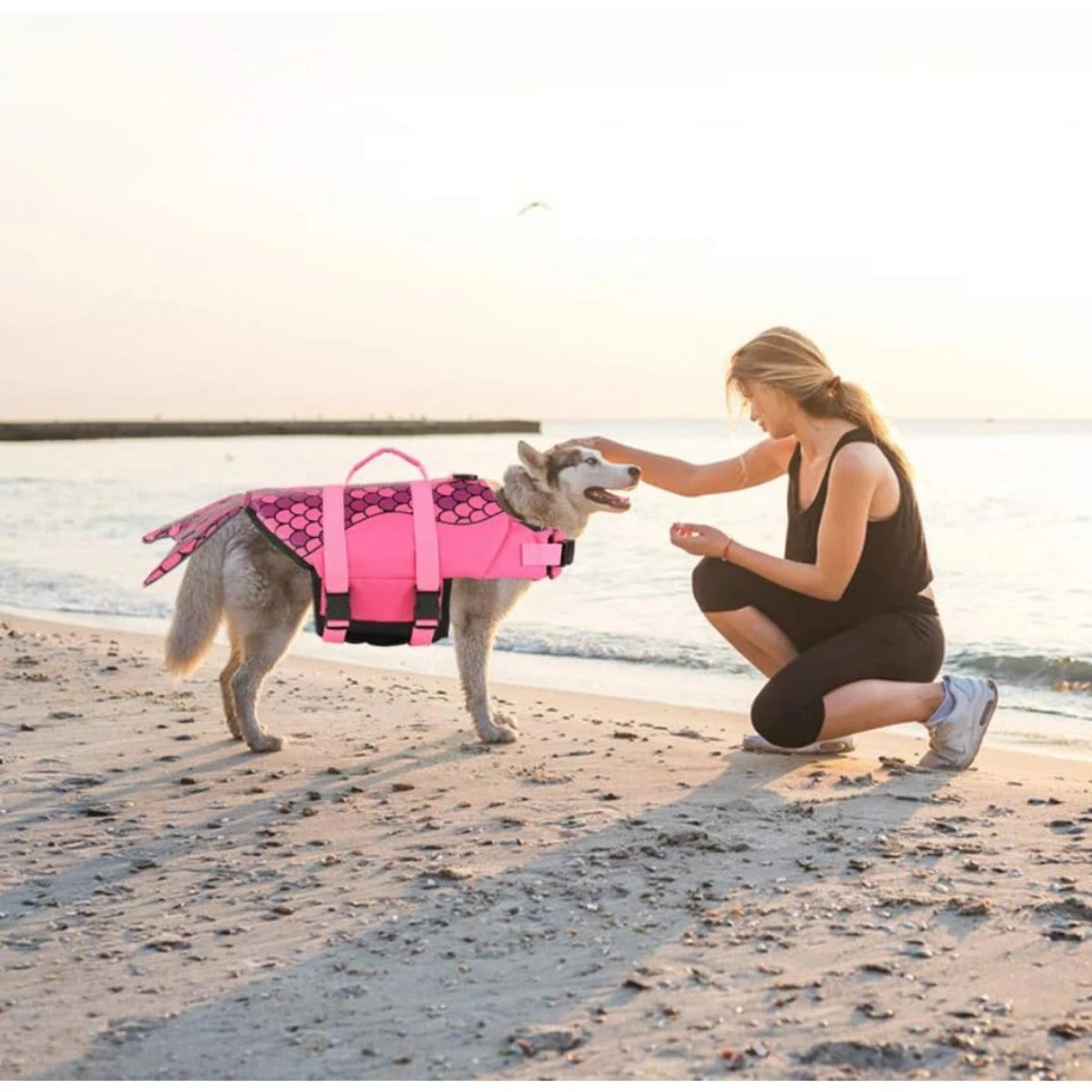 Eine Frau hält einen Hund mit einem rosa Meerjungfrauen-Schwimmweste am Strand bei Sonnenuntergang.