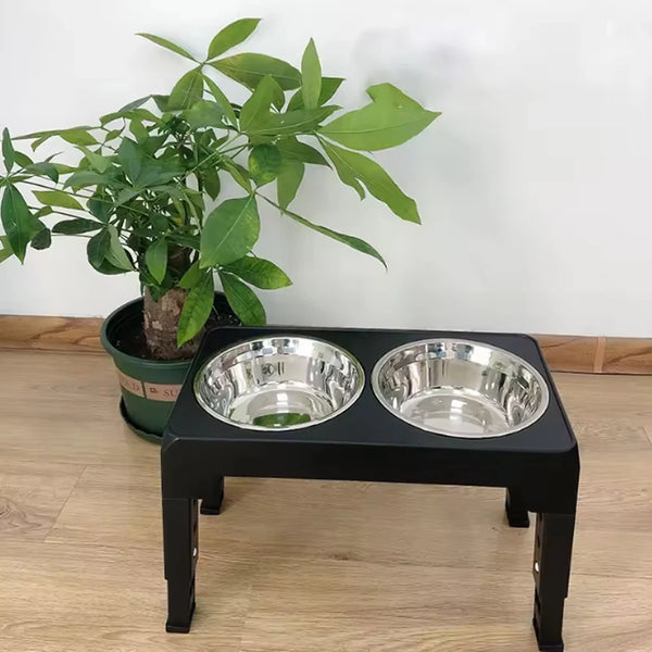 A black adjustable raised dog feeder with two stainless steel bowls sits on a wooden floor next to a potted green plant.