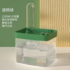 A clear glass water fountain with a green base and an attached metal filter, set against a light wood background.