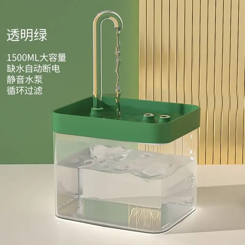 A clear glass water fountain with a green base and an attached metal filter, set against a light wood background.