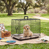A black foldable metal pet crate with a security lock sits on a woven mat in a grassy outdoor setting.