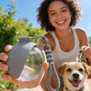 A young woman holds a gray portable pet water bottle with a clear dispenser and lanyard, smiling while a dog looks at the cam