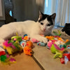 A white and black cat is lying next to a colorful bundle of mixed cat play toys.