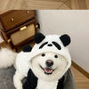 A white dog wearing a black and white panda-themed fleece winter hood coat sitting on a gray surface.
