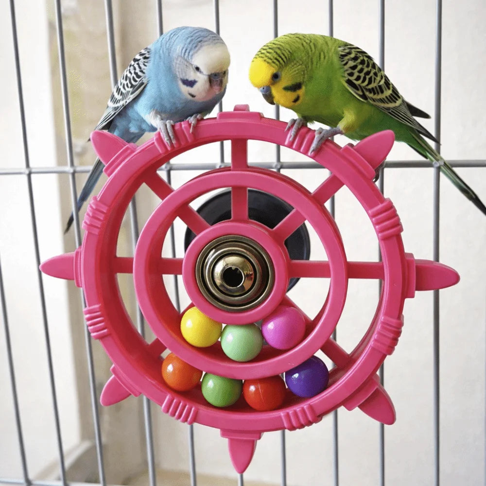 Two small parrots are perched on a pink interactive spinner rattle toy inside a wire cage.