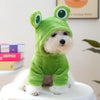 A small dog is dressed in a green fabric hoodie resembling a frog, sitting on a white surface.