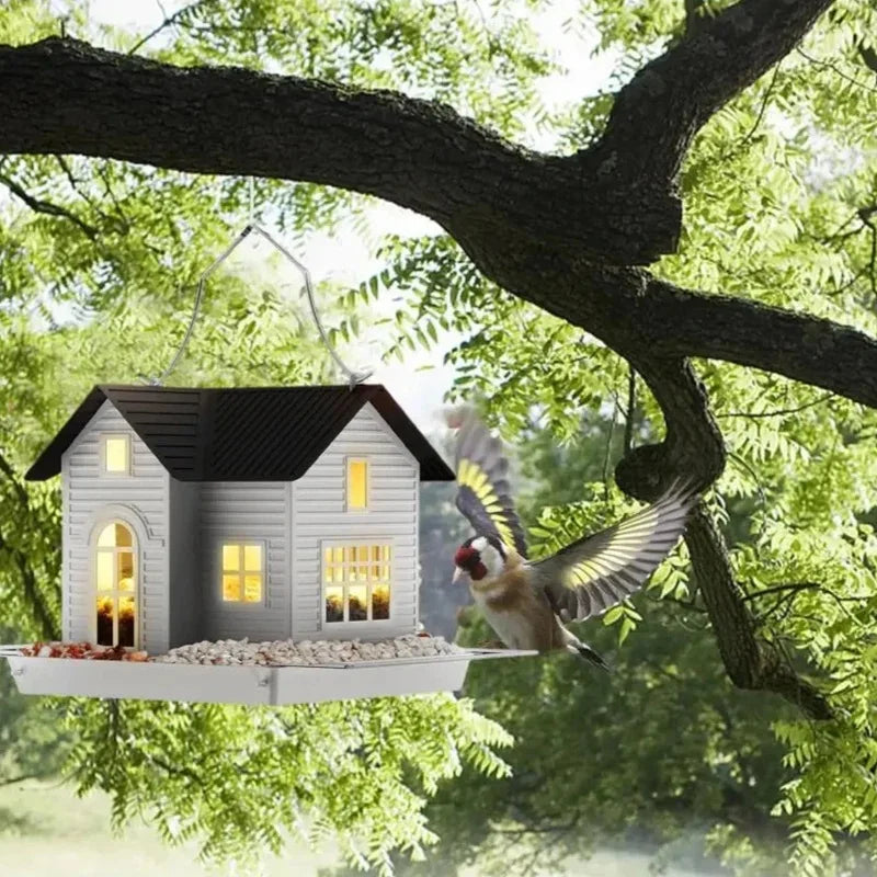 A solar outdoor bird feeder lantern is hanging from a tree branch, illuminating a small wooden house structure.