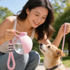 A woman is holding a pink portable pet water bottle with a lanyard, interacting with a golden dog outdoors.