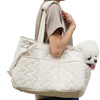 A person is holding a cream-colored quilted portable pet carrier bag with a handle, featuring a small white dog peeking out.