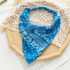 Una bandana azul brillante con estampado floral y paisley blanco, exhibida sobre una tabla de madera beige junto a un tejido 