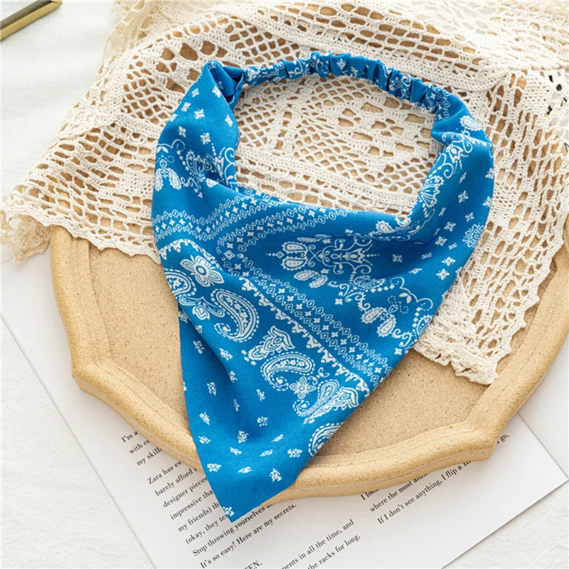 Una bandana azul brillante con estampado floral y paisley blanco, exhibida sobre una tabla de madera beige junto a un tejido 