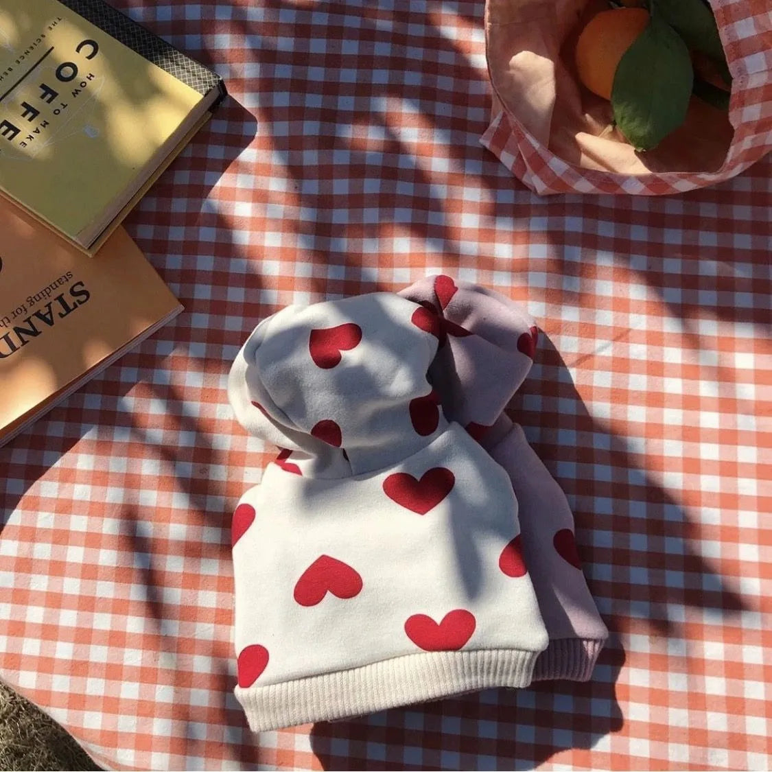Sudadera de peluche blanca y roja con estampado de corazón, doblada sobre un mantel a cuadros junto a un libro y fruta.