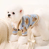 Un perro blanco con suéter azul estampado de oso marrón, sentado en una manta color crema.