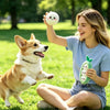 A young woman is smiling while holding a green plush sausage dog chew toy and interacting with a happy dog in a grassy park.