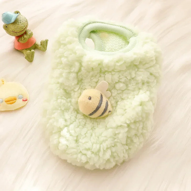 A light green fuzzy pet sweater with a small bee appliqué rests on a white surface next to a small green animal and a yellow 