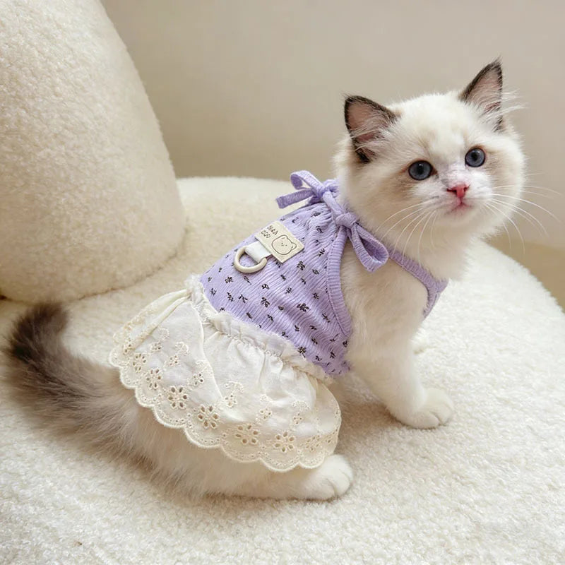 A small white and blue cat wearing a lavender floral print dress with thin straps, sitting on a white fluffy surface.