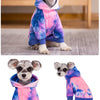 A small dog wearing a tie-dye fleece hoodie in shades of pink, blue, and purple.