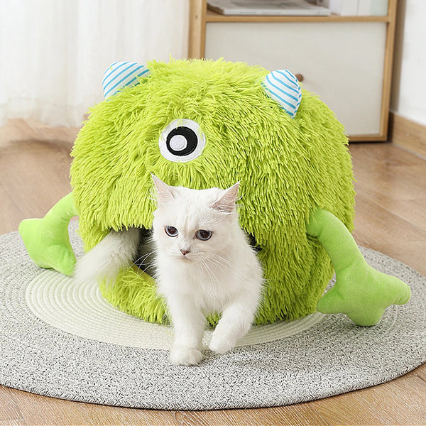 A small white kitten is peeking out from a bright green plush monster cave bed.