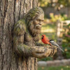 A realistic resin sculpture of a bearded man hugging a tree trunk, with a small bird perched on his hand.