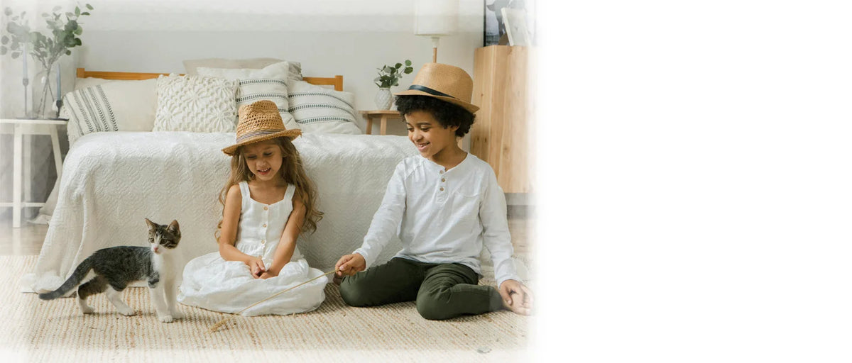A young boy and girl sitting on a light-colored rug with a cat next to them in a bright bedroom.