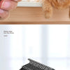 A hand holds a double-sided dog grooming comb and brush set, with a cat looking at the product.